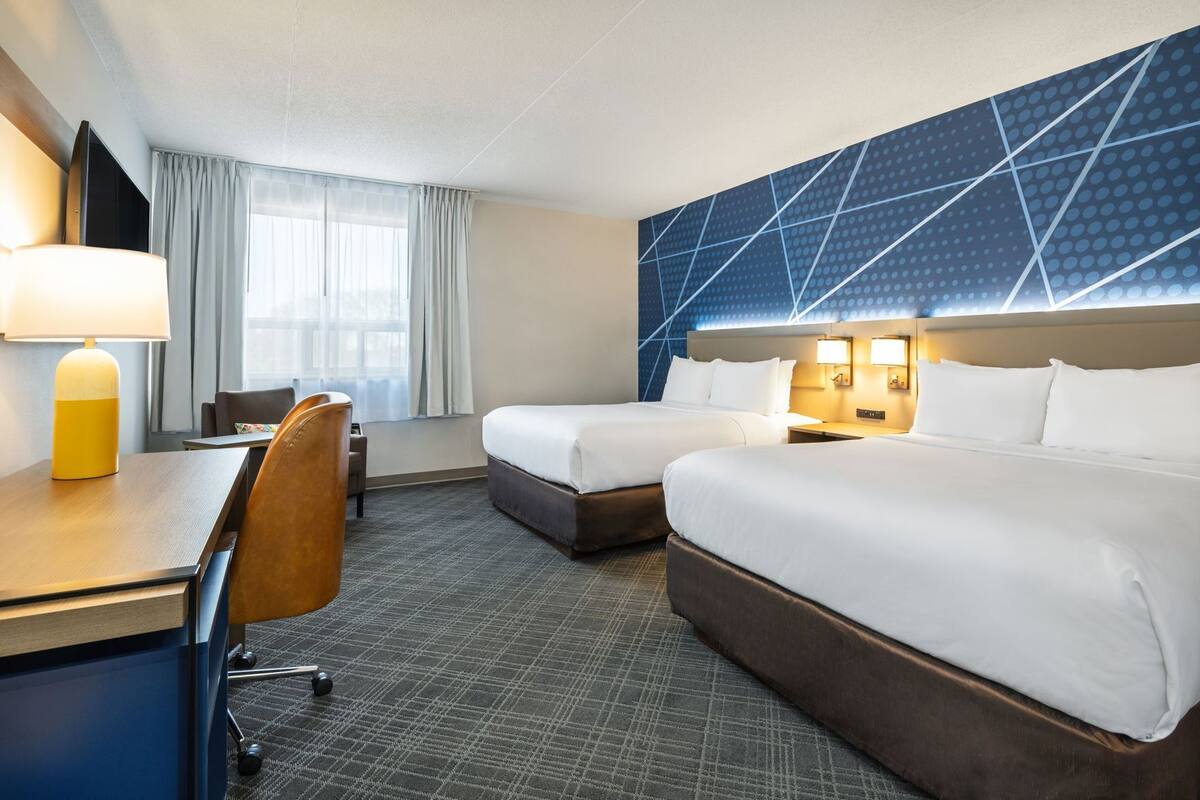 A hotel room with two beds, a desk, a chair, a TV, and a large window with curtains.