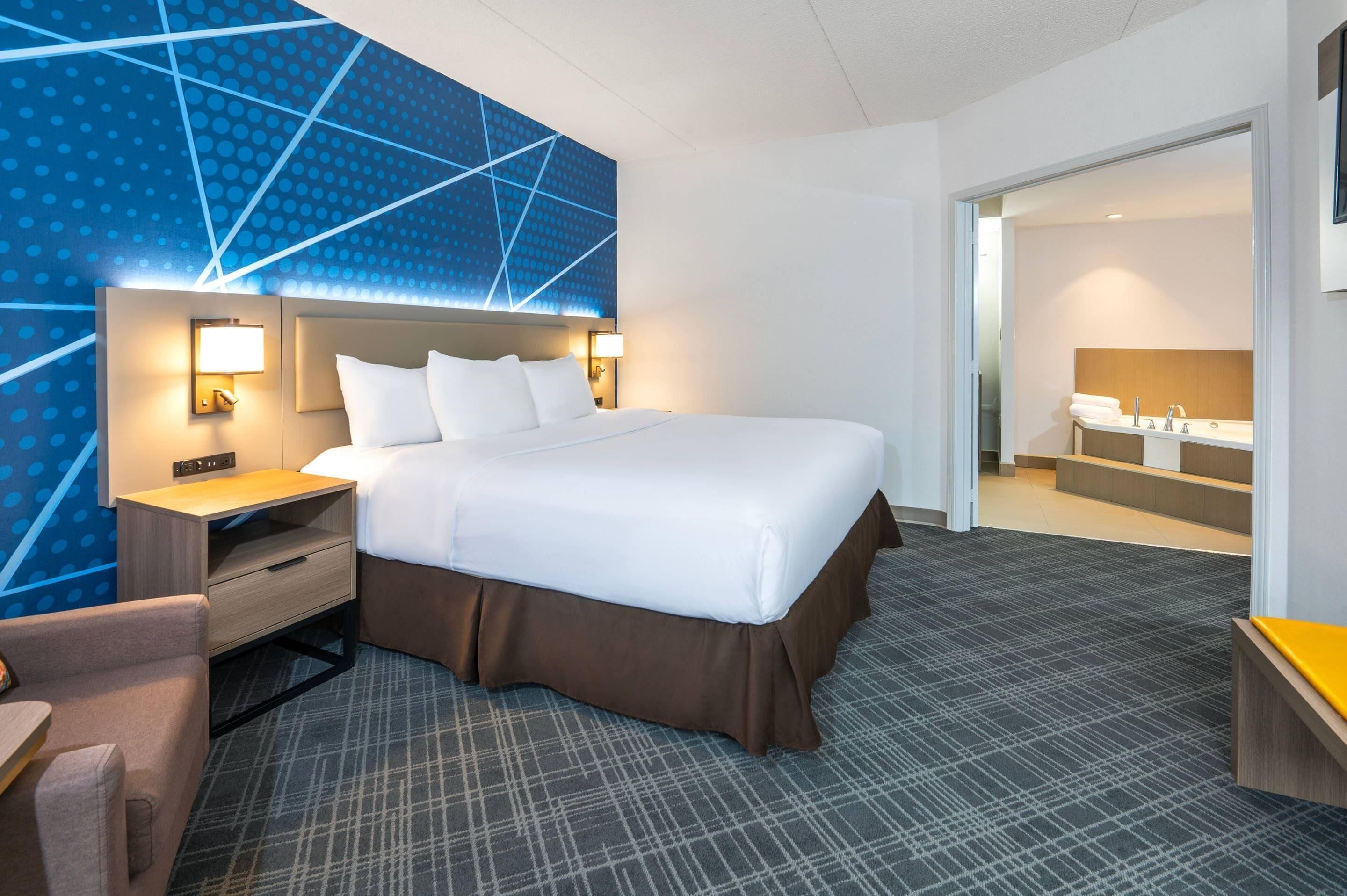 A hotel room with a large bed, bedside tables, a desk, and a bathroom with a sink and mirror.