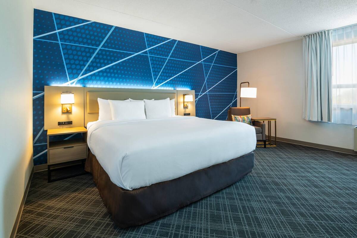 A hotel room with a large bed, two bedside tables with lamps, a small table with a chair, and a window with curtains.