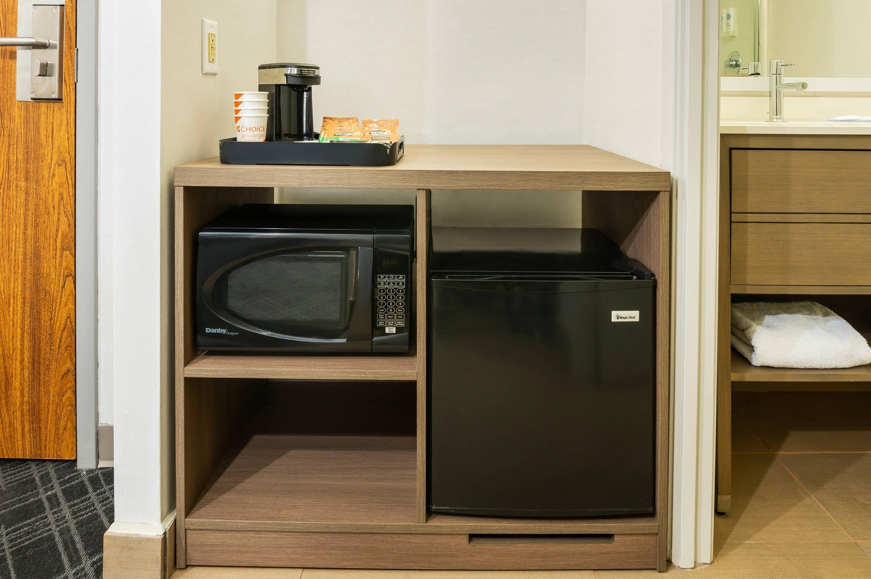 Una kitchenette compacta con microondas, refrigerador, cafetera y espacio para artículos de aseo.