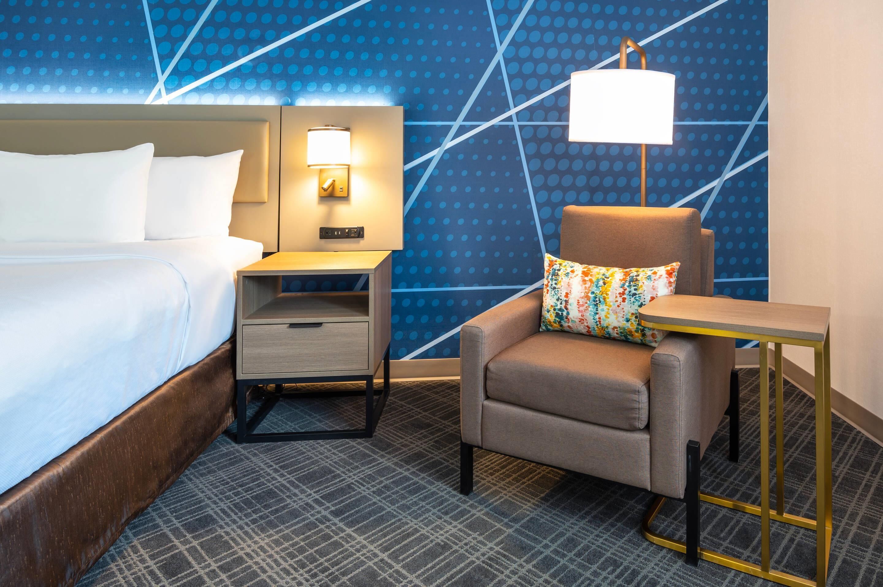 Ein Hotelzimmer mit einem Bett, einem Nachttisch mit Lampe, einem Sessel mit farbigem Kissen und einem kleinen Tisch ebenfalls mit Lampe.