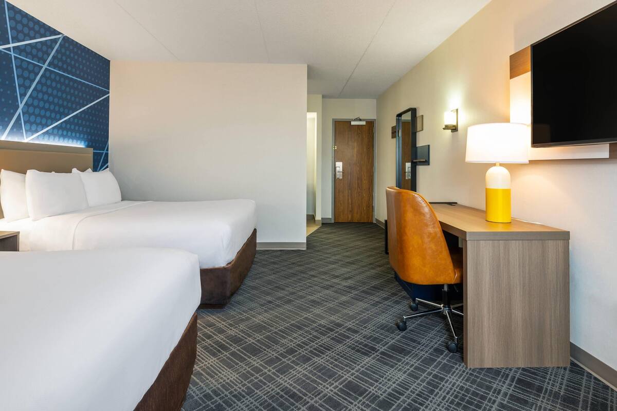 A hotel room with two beds, a desk, a chair, a TV, and a lamp.