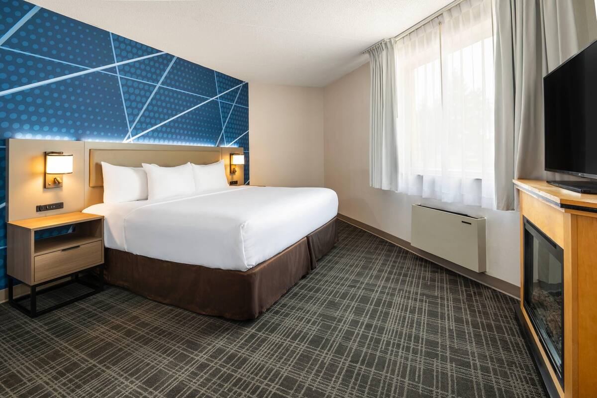 A hotel room with a large bed, bedside lamps, a flat-screen TV, a window with curtains, and a patterned carpet.