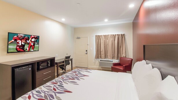 A hotel room with a bed, a desk with a chair, a television mounted on the wall, and a window with curtains.