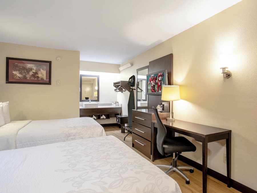 A hotel room with two beds, a desk, a chair, a mirror, a television, and a lamp.