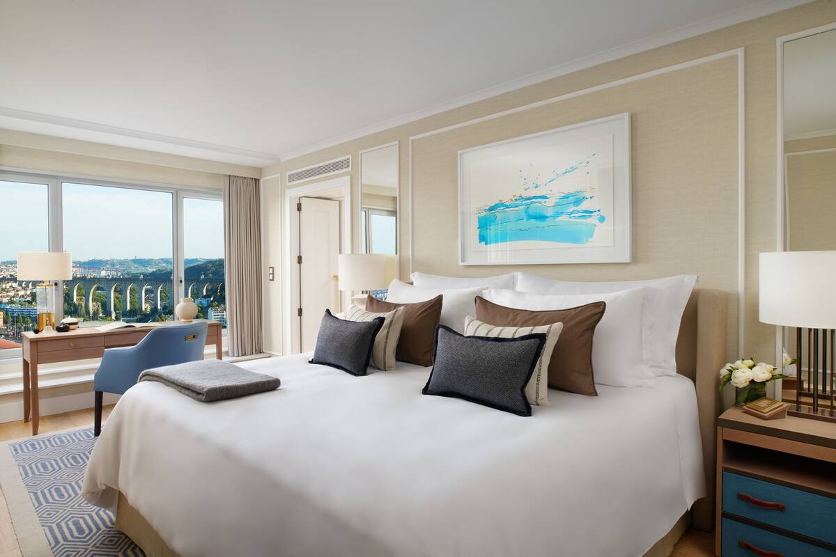 A hotel room with a large bed, a desk, and a chair. There is a window with a view of a bridge and buildings.
