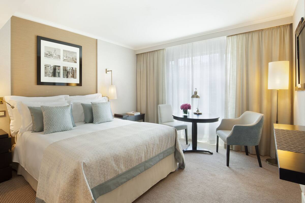 A hotel room with a large bed, two bedside tables, a desk with a chair, a large window with curtains, and a framed picture on the wall.