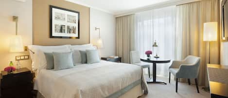 A hotel room with a large bed, two bedside tables, a desk with a chair, a large window with curtains, and a framed picture on the wall.