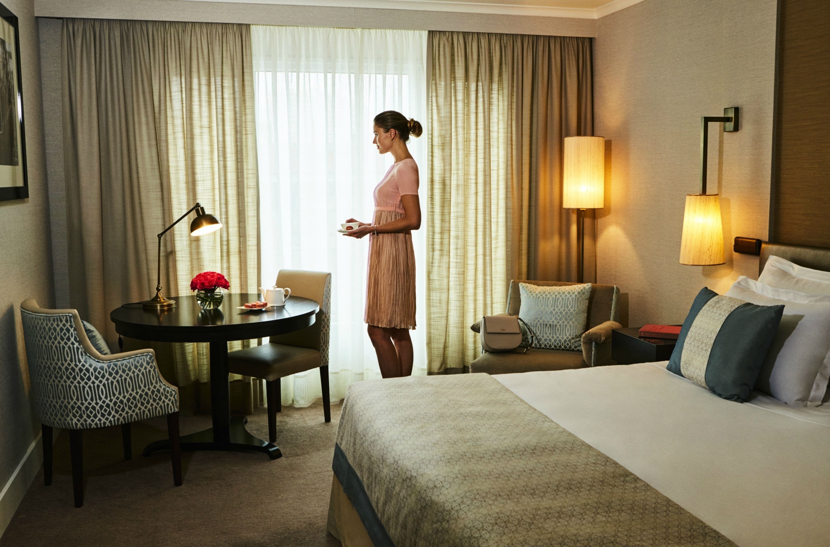 A hotel room with a bed, a round table with a lamp, a chair, and a small table with a vase of flowers.