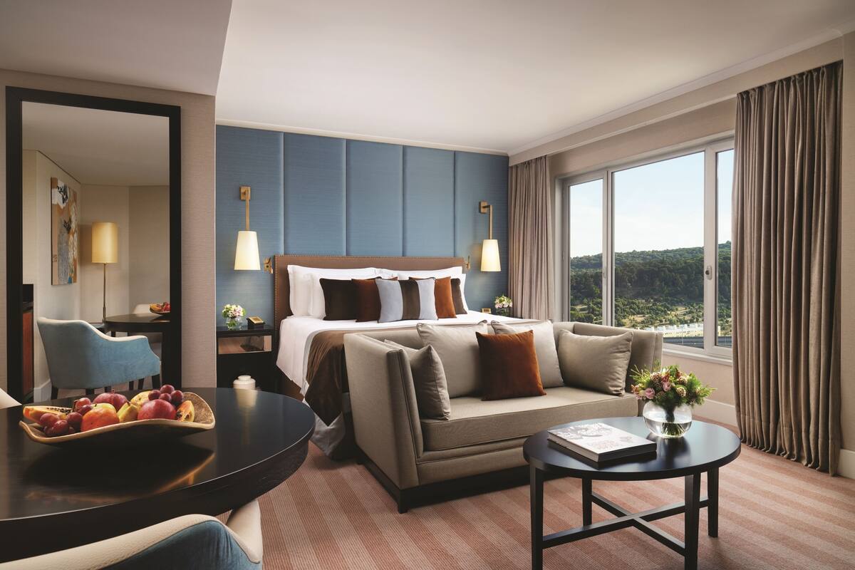 A hotel room with a bed, a sofa, a dining table with fruit, a vase with flowers, and a view of the outdoors.