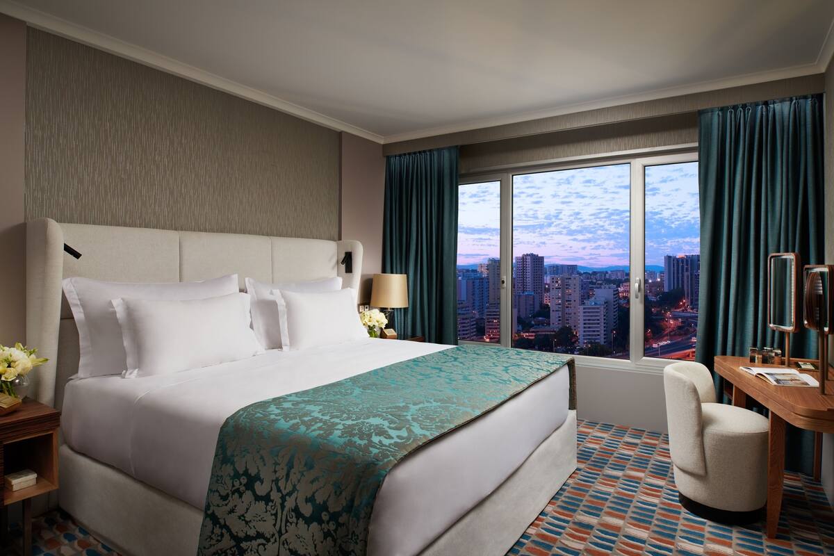A hotel room with a large bed, a desk, and a view of the cityscape through the window.