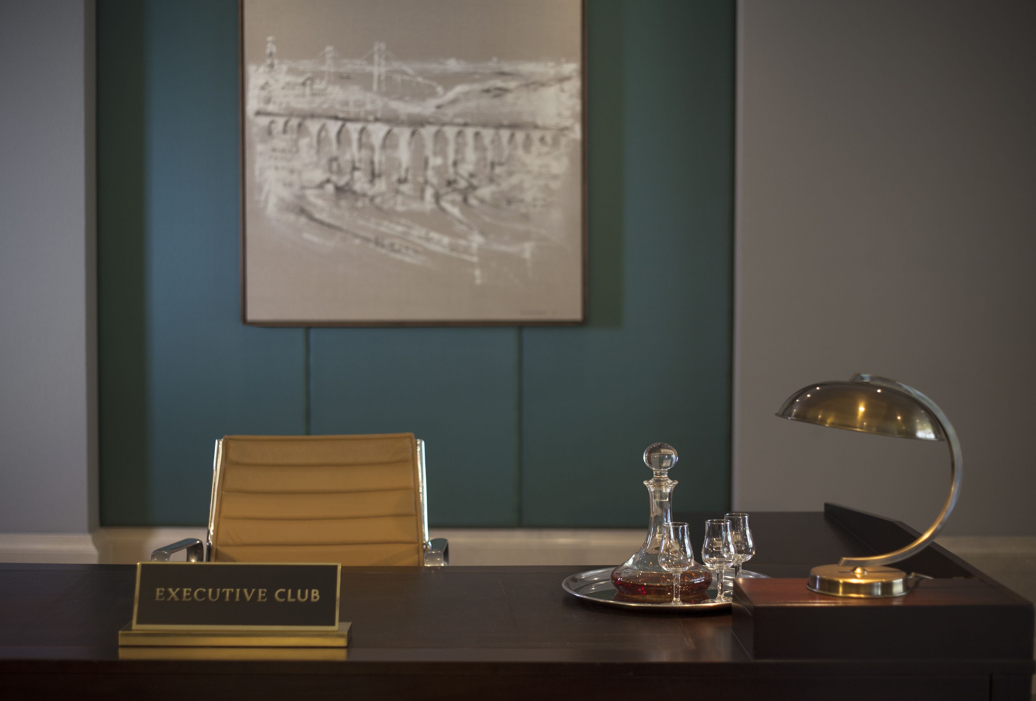 Habitación de hotel con un escritorio de madera oscura, una placa con la inscripción “EXECUTIVE CLUB”, un decantador de vidrio, dos copas de vino y una lámpara moderna.