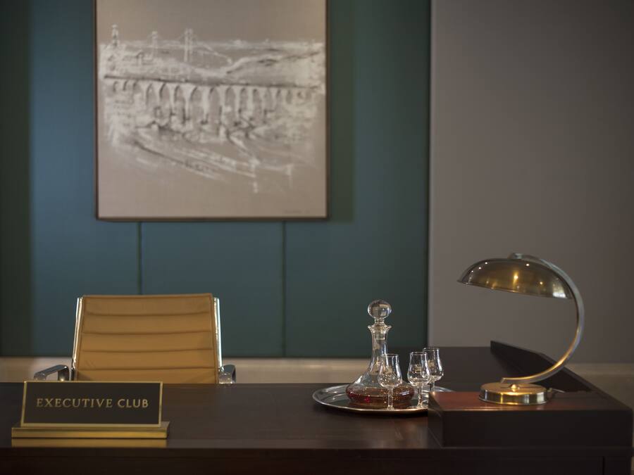 Habitación de hotel con un escritorio de madera oscura, una placa con la inscripción “EXECUTIVE CLUB”, un decantador de vidrio, dos copas de vino y una lámpara moderna.