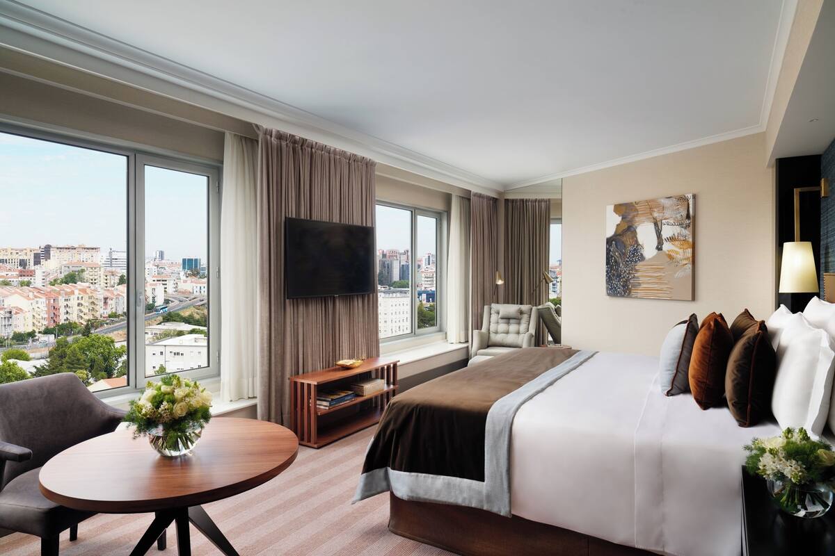 A hotel room with a large bed, a TV, a desk with a chair, and a view of the city.
