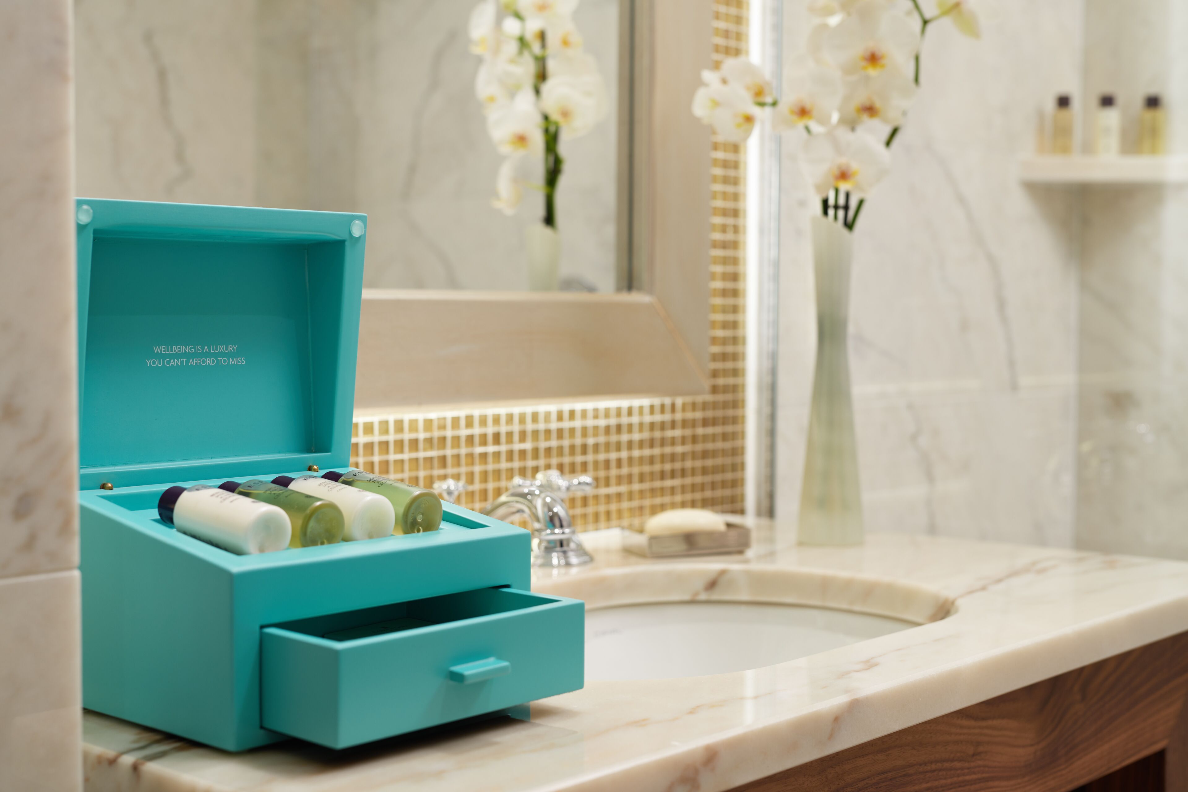 Une salle de bain avec une boîte turquoise contenant des articles de toilette, un lavabo et un miroir orné d’orchidées.
