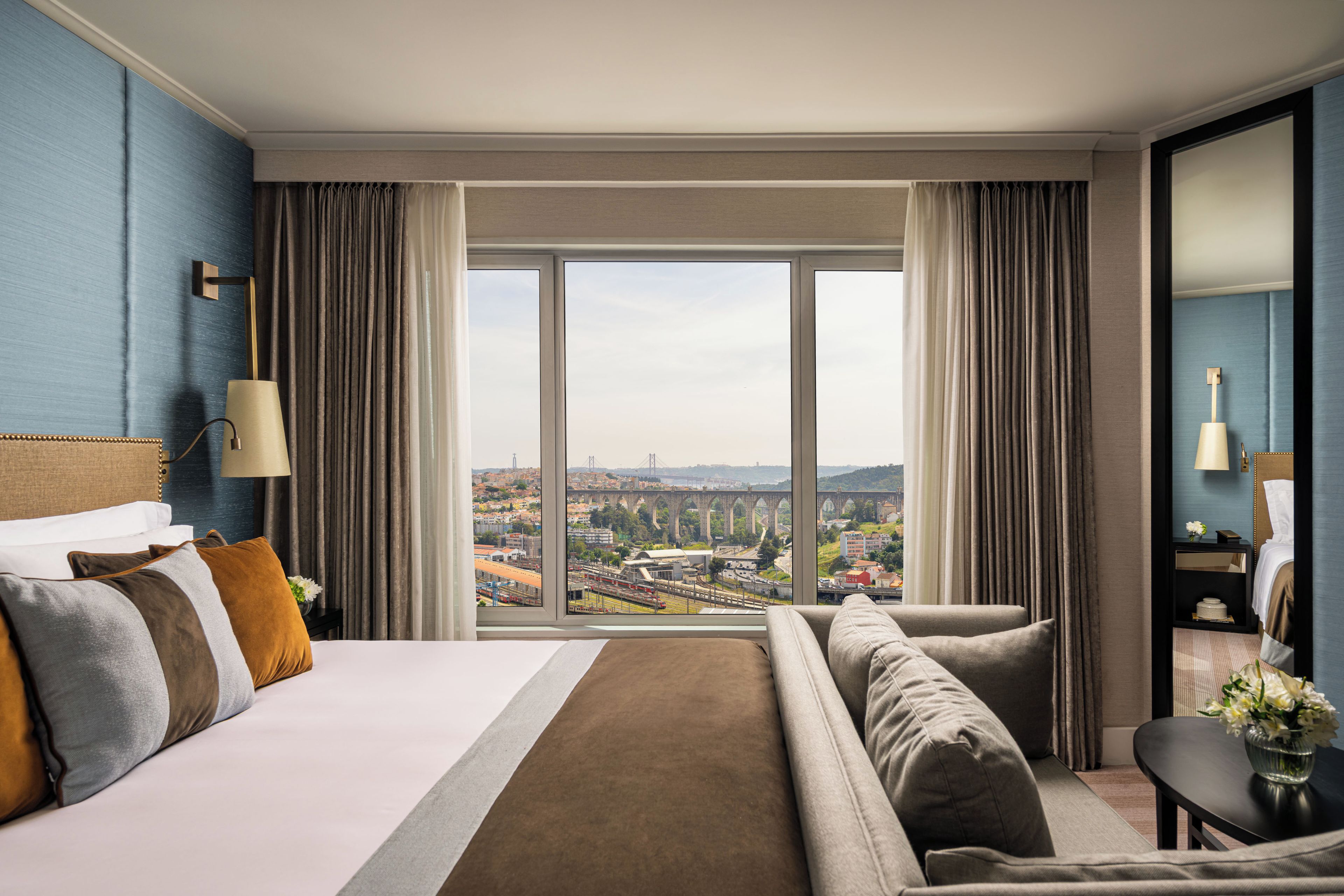 A hotel room with a large bed, a sofa, a view of a cityscape, and a nightstand with a lamp.