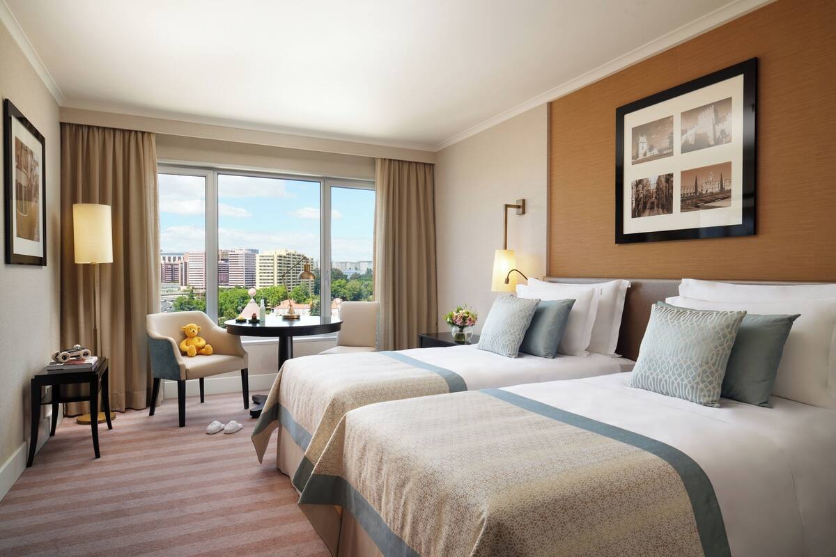 A hotel room with two beds, a desk, a chair, and a large window offering a city view.