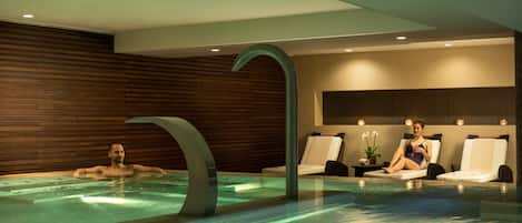 Una piscina interior moderna con un grifo curvo distintivo, paneles de madera en las paredes y dos sillones de descanso.