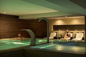 A modern indoor pool with a unique curved faucet, wooden paneling on the walls, and two lounge chairs.