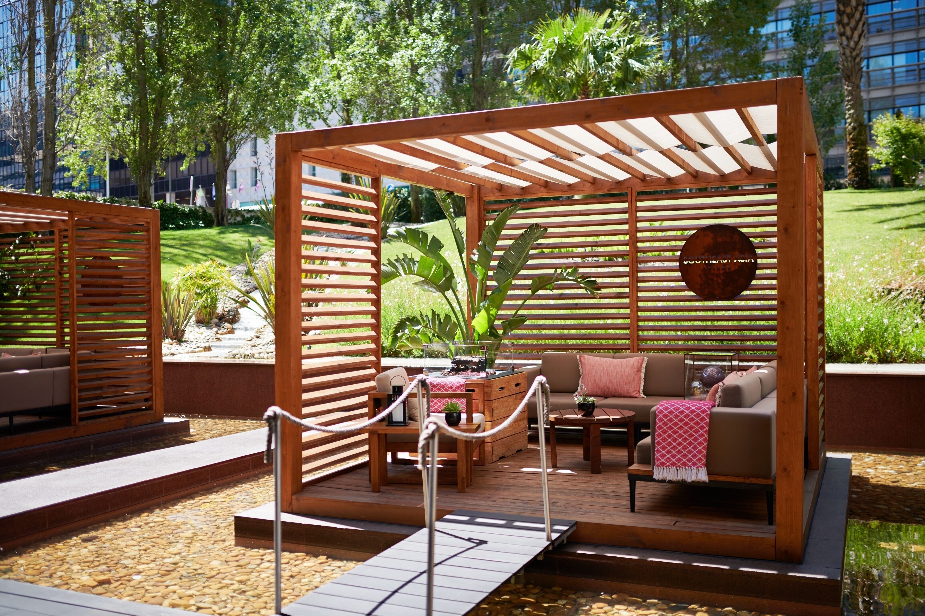 Une pergola en bois avec un espace salon comprenant un canapé et une table basse, le tout entouré d’un jardin agrémenté de plantes et d’un petit élément décoratif aquatique.