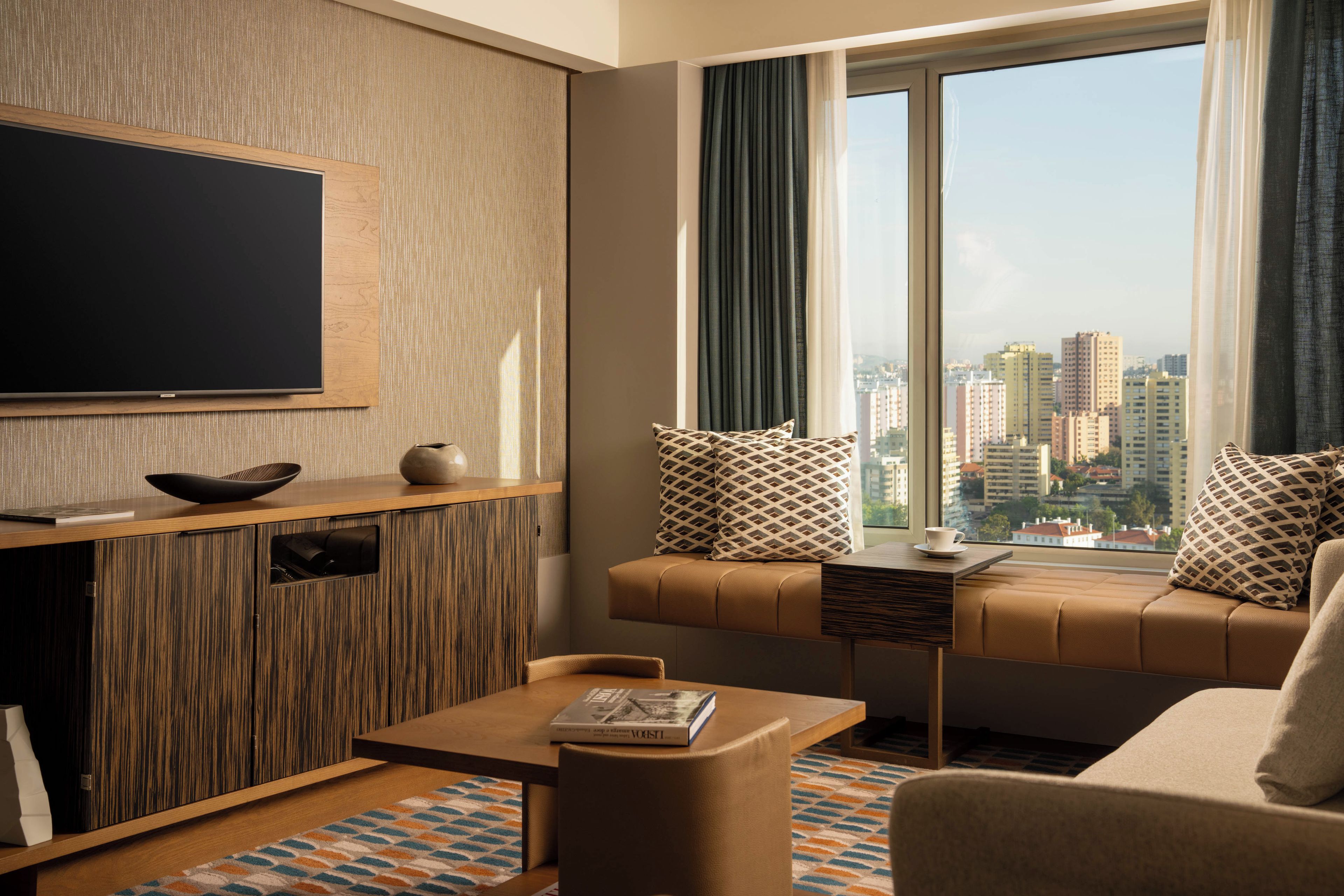 A modern living room with a flat-screen TV, a sofa with patterned cushions, a coffee table with a book, and a view of a cityscape through large windows.