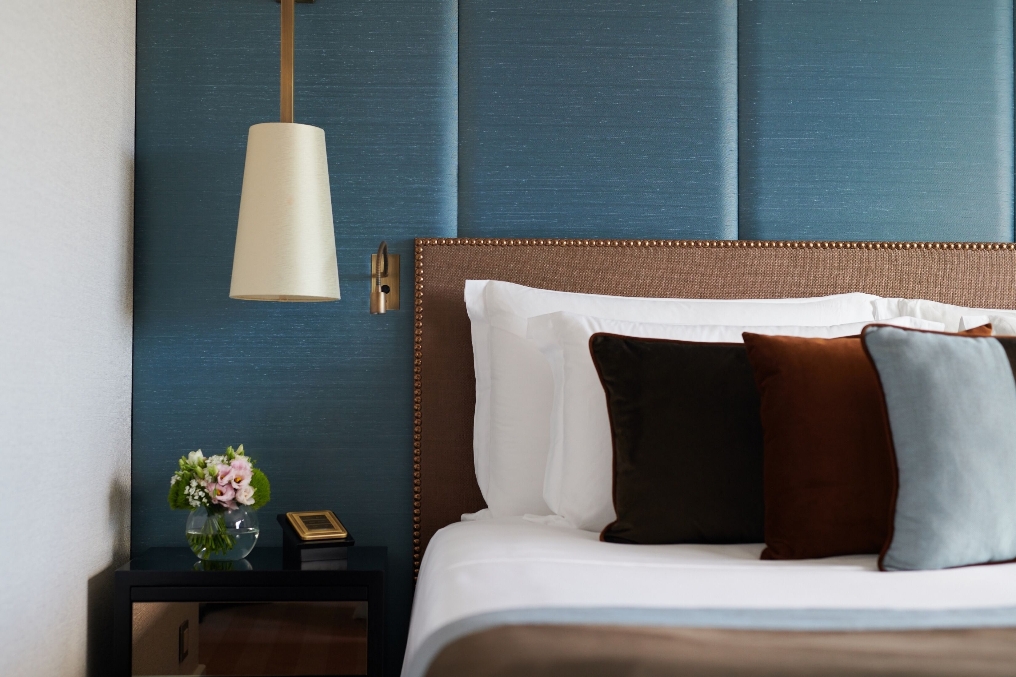 Een keurig opgemaakt bed met een bruin hoofdbord, witte en bruine kussens, en een nachtkastje met een bloevas en een klein decoratief object.