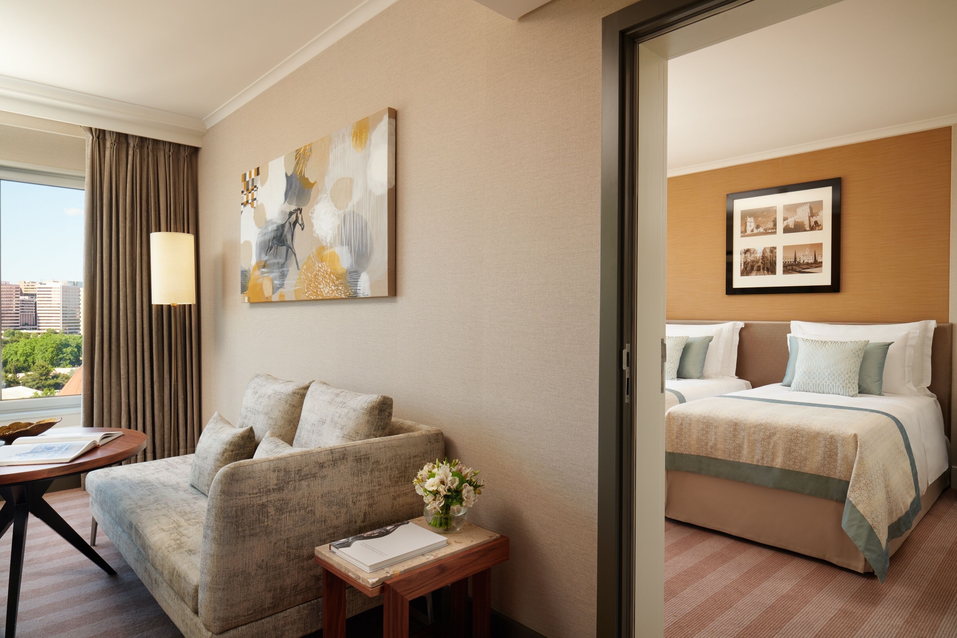 Una camera d'albergo con divano, un piccolo tavolo con un vaso di fiori, un letto e una cornice con un quadro al muro, oltre a vista sulla città dalla finestra.