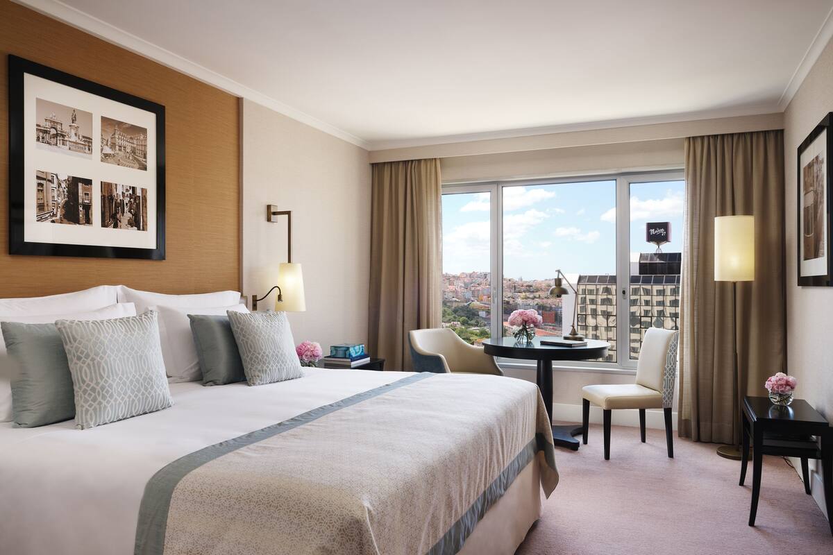 A hotel room with a large bed, a desk with a chair, a view of the city, and a framed photo on the wall.