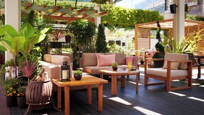 Un patio al aire libre con mobiliario de madera, plantas en macetas y una estructura de pérgola.