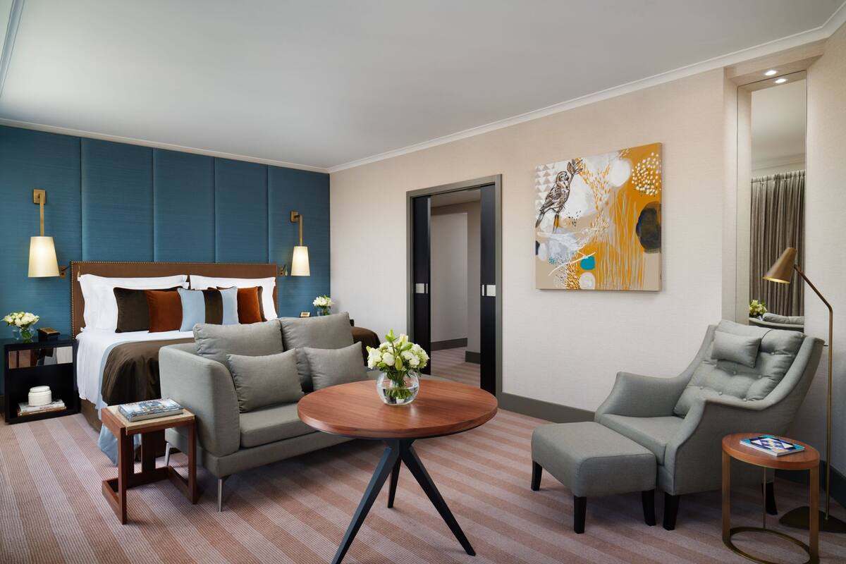 A hotel room with a bed, a sofa, a round coffee table, and a chair. There is a painting on the wall and a lamp on a side table.