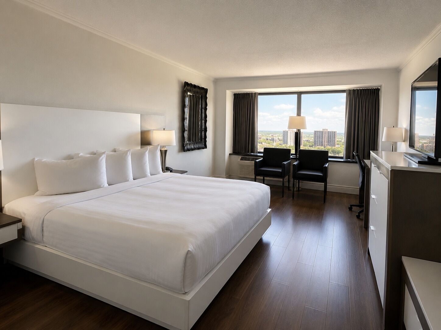 A hotel room with a large bed, a desk, two chairs, a television, and a window with a city view.