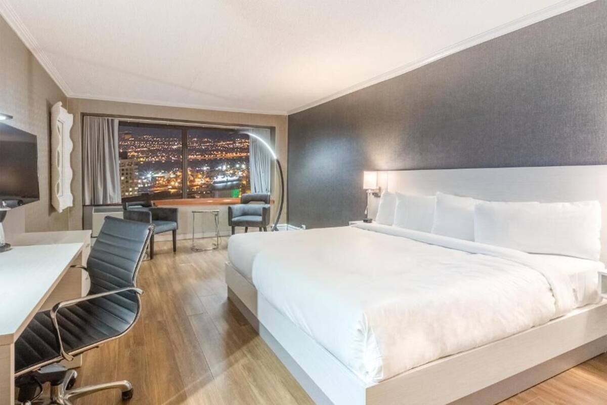 A hotel room with a large bed, a desk with a chair, a TV, and a view of a cityscape at night.