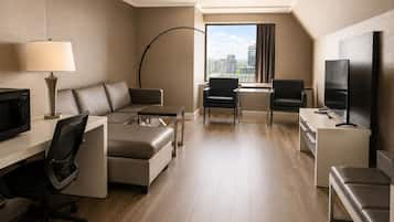 A modern hotel room with a sofa, two chairs, a desk with a lamp, a flat-screen TV, and a large window offering a city view.