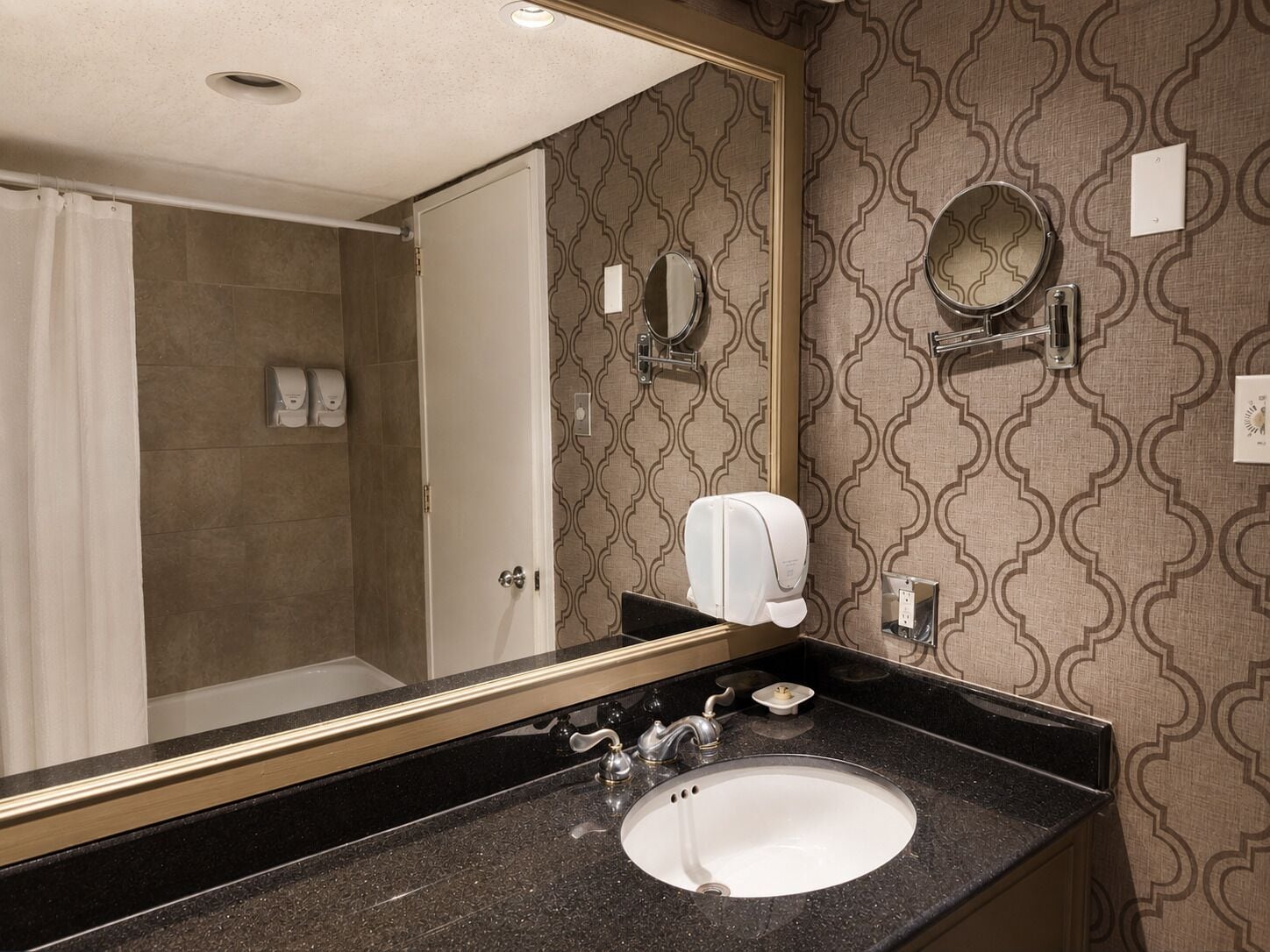 A hotel bathroom with a sink, mirror, towel dispenser, and a shower area.
