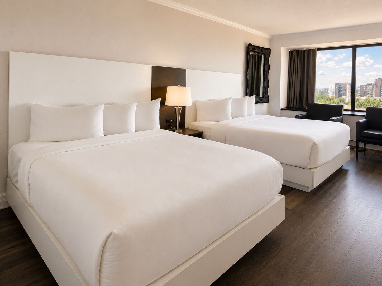 Een hotelkamer met twee bedden, een bureau, een stoel, een lamp en uitzicht op de stad.