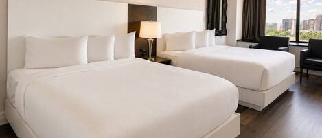 Quarto de hotel com duas camas, uma escrivaninha, uma cadeira, um abajur e vista para a cidade.
