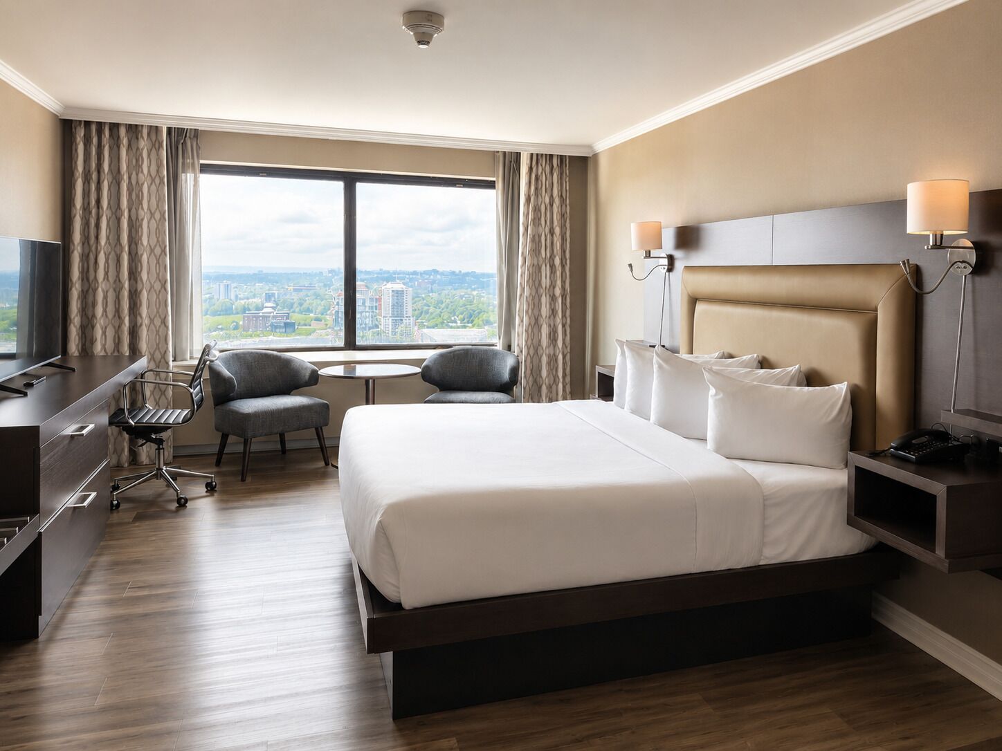 Habitación de hotel con una cama grande, un escritorio, una silla, un televisor y vistas a la ciudad.