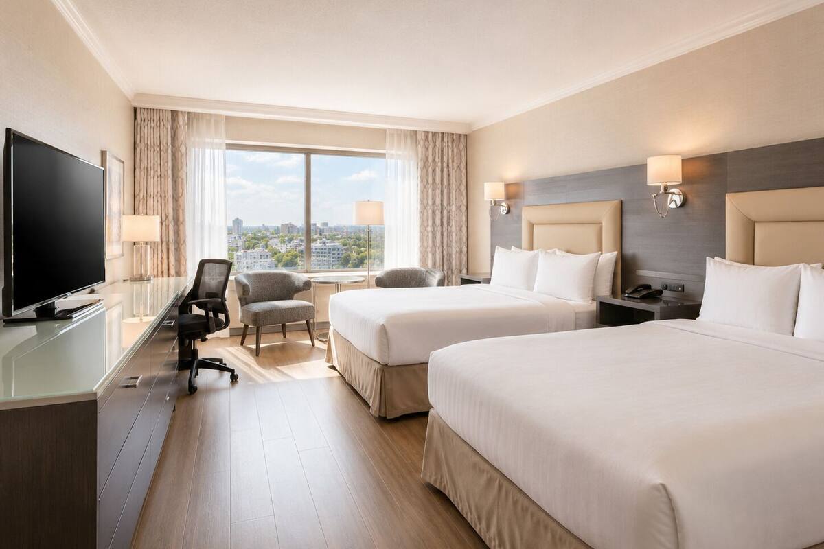 A hotel room with two beds, a desk with a TV, a chair, a lamp, and a view of the city.