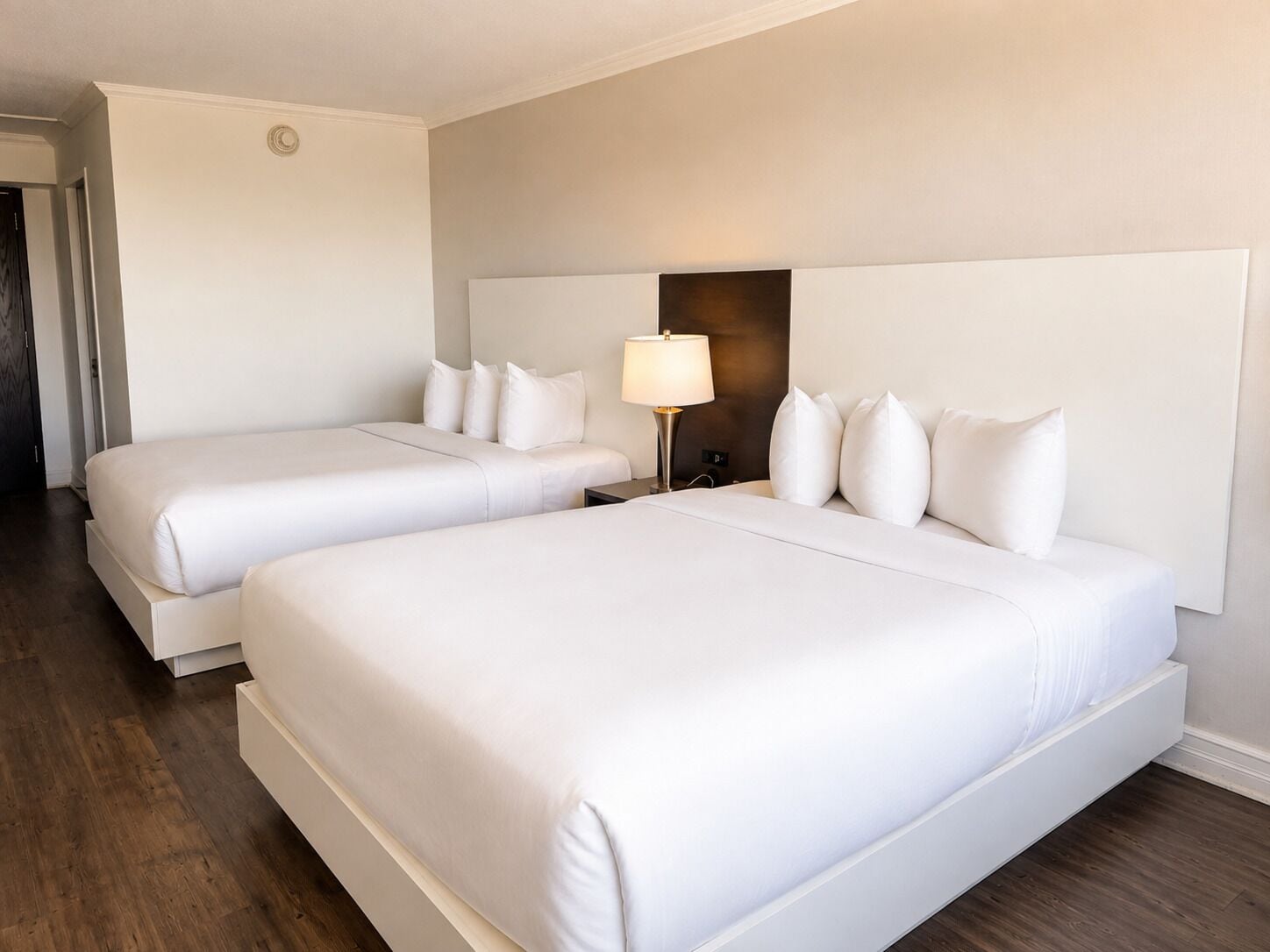 A hotel room with two beds, a nightstand with a lamp, and a wall-mounted light fixture.