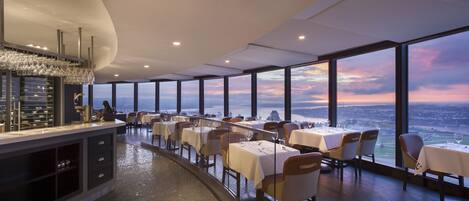 Un restaurante con vistas panorámicas de un paisaje urbano y un río, que presenta un interior moderno con mesas y sillas dispuestas para comer.