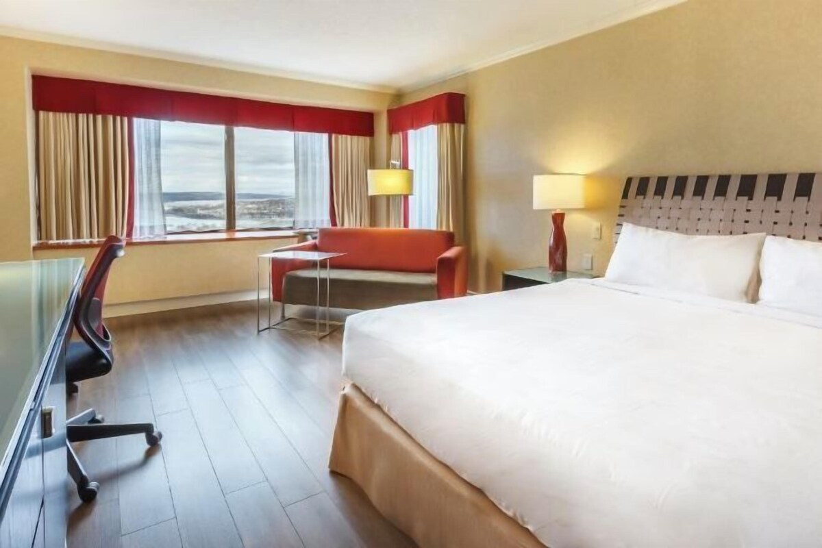 A hotel room with a bed, a desk, a chair, a window with curtains, and a lamp.