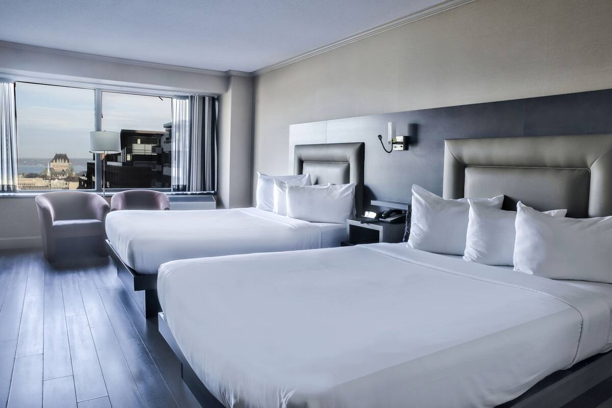 A hotel room with two beds, a desk, a chair, and a large window with a view of buildings.