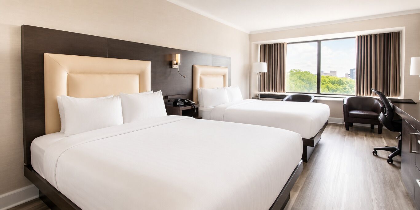 A hotel room with two beds, a desk, a chair, and a large window.