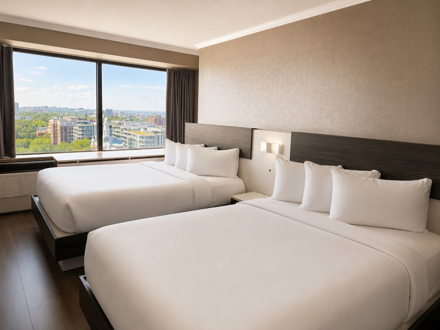 Une chambre d’hôtel avec deux lits, une grande fenêtre donnant sur la ville et un luminaire fixé au mur.