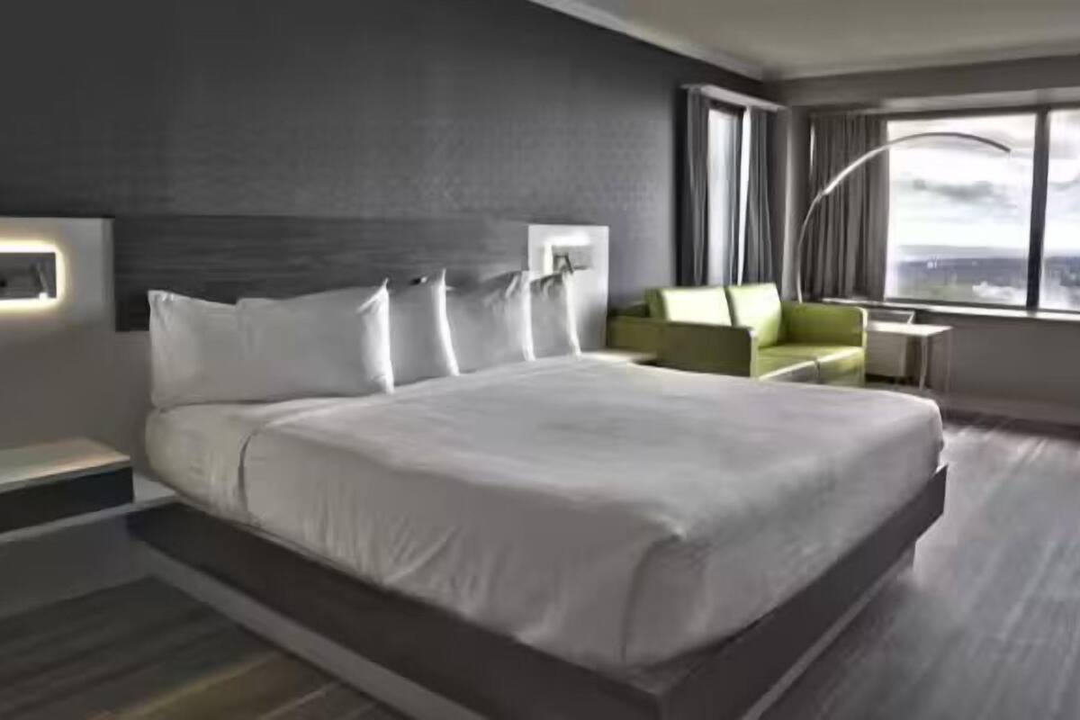 A modern hotel room with a large bed, a green armchair, a desk, and a view of the city through large windows.