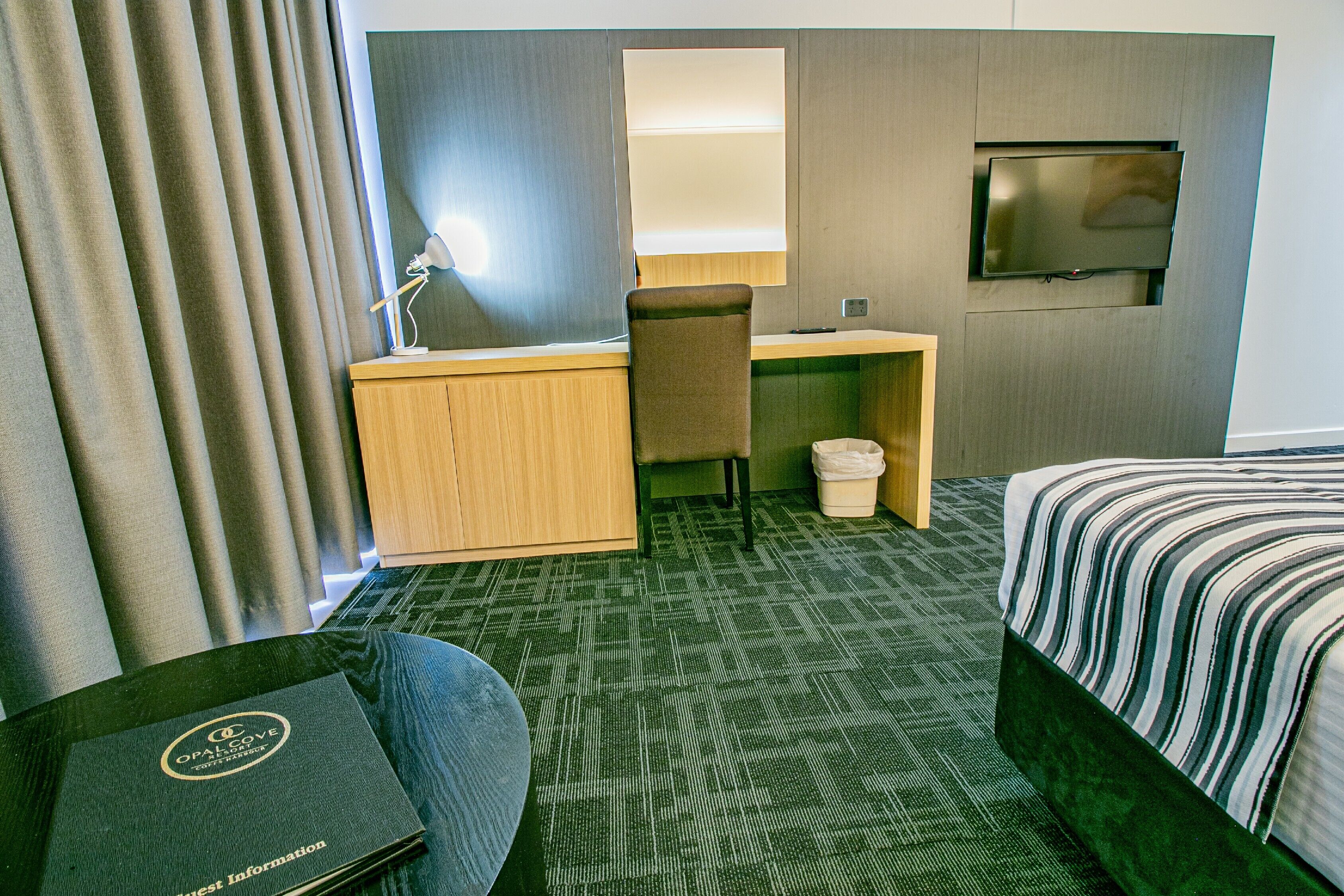 A hotel room with a bed, a desk, a chair, a TV, and a small trash bin.