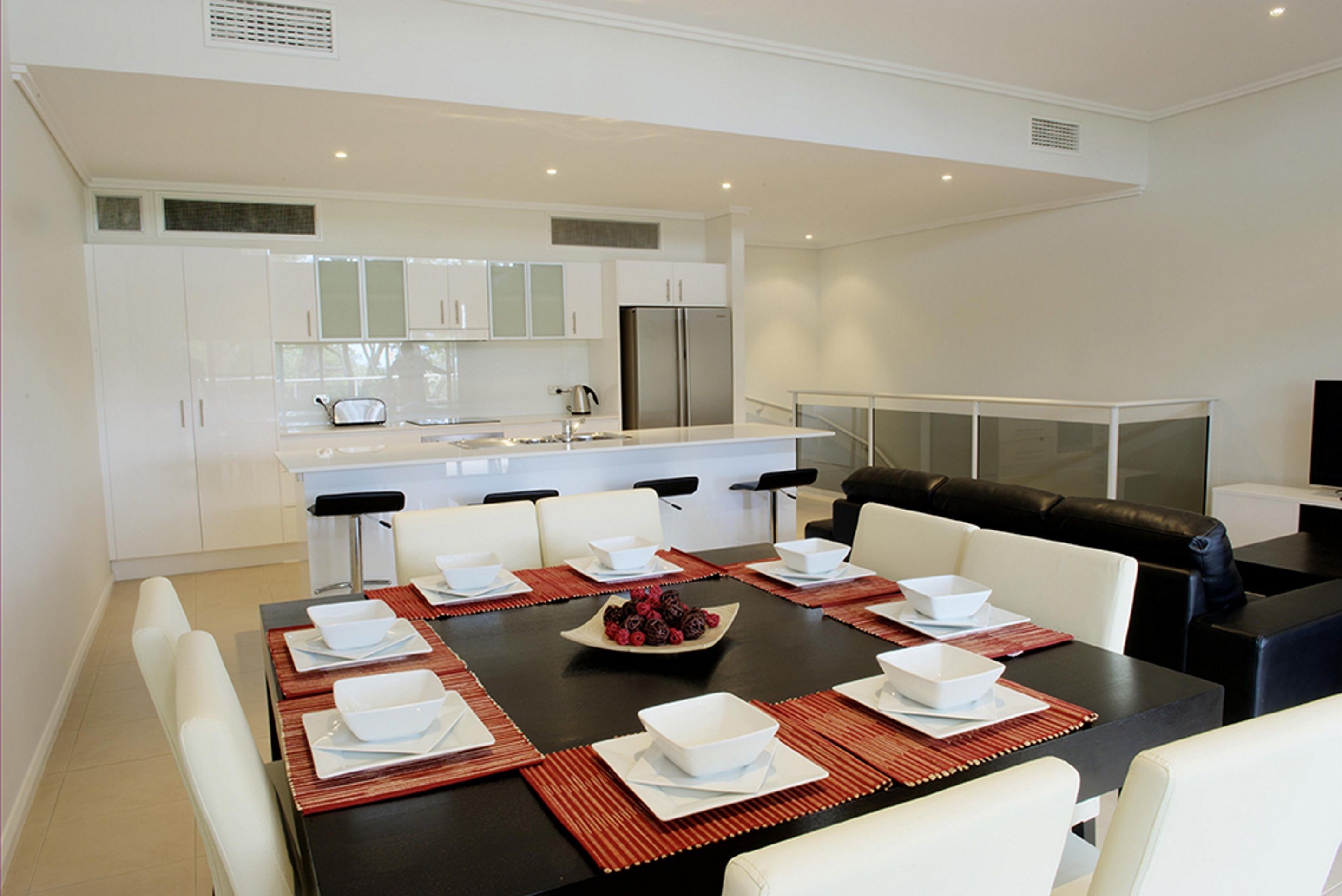 Une cuisine moderne avec des meubles blancs, un réfrigérateur en acier inoxydable et un coin repas comprenant une table noire et des chaises blanches.