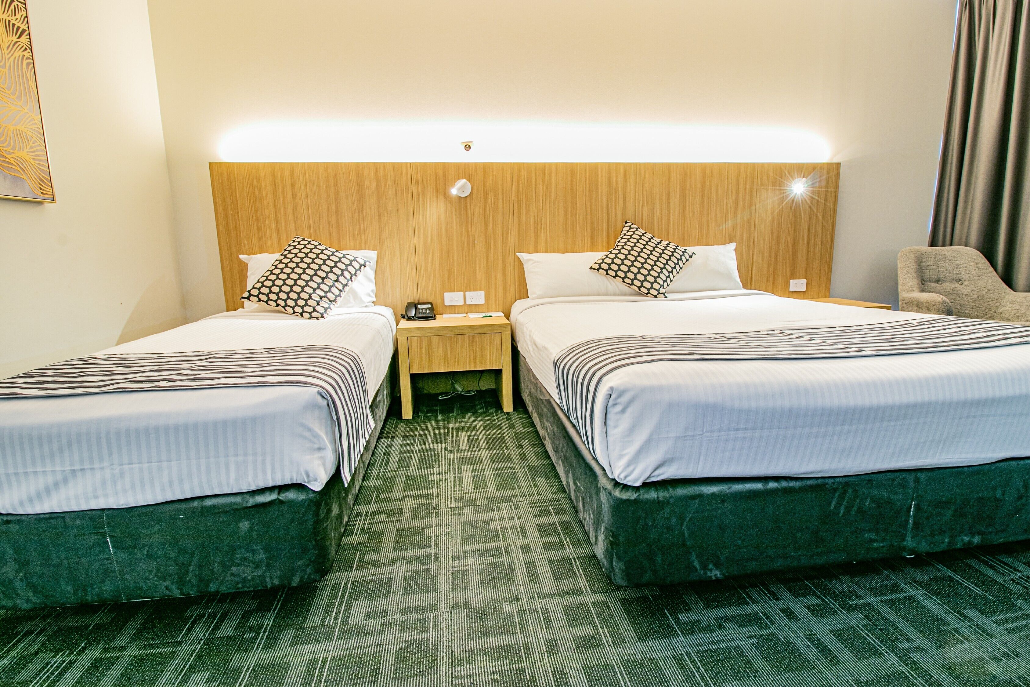A hotel room with two beds, a wooden headboard, a nightstand with a phone, and a green carpet.