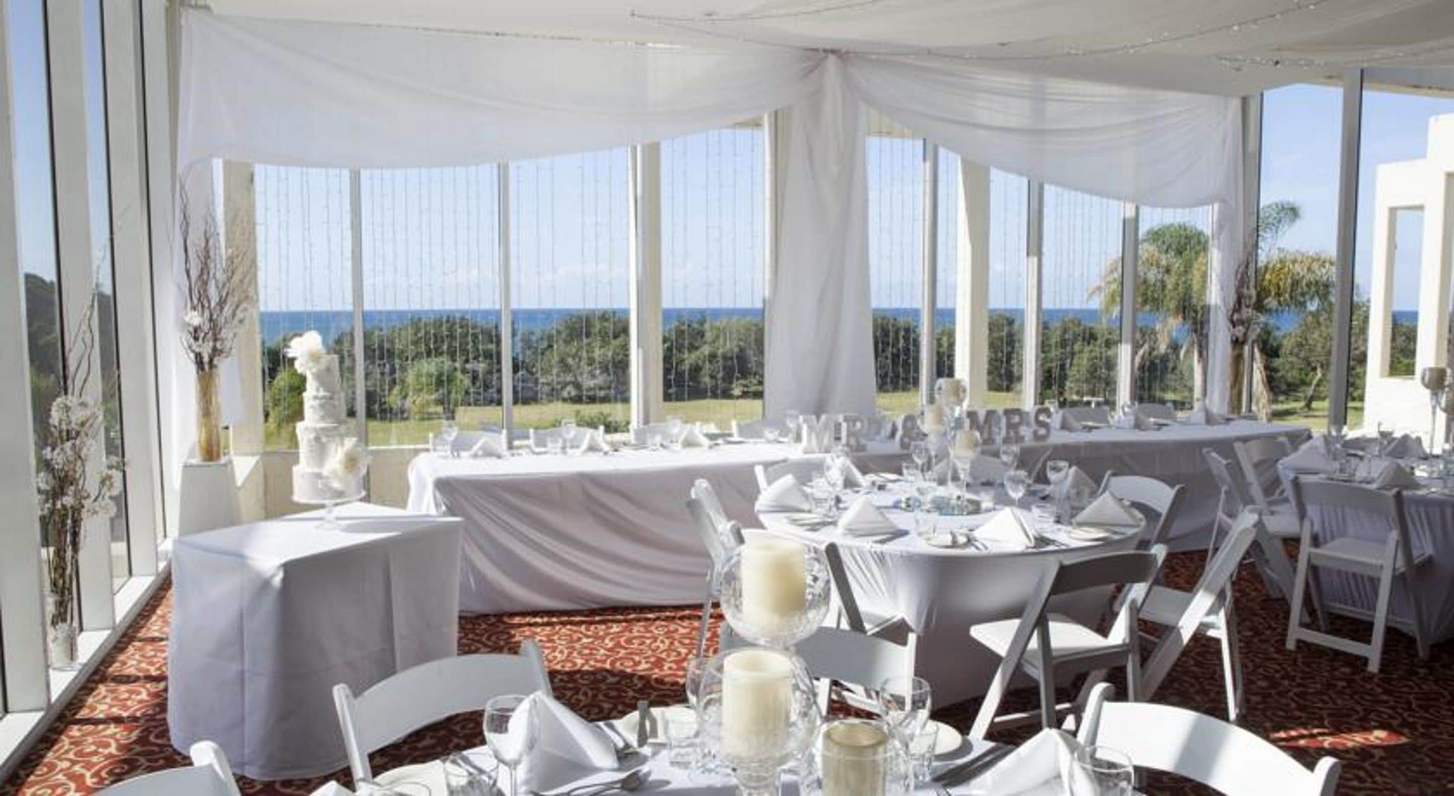 Una sala da banchetto con tavoli rotondi apparecchiati per un evento formale, ampie finestre che offrono vista sull'esterno e un soffitto drappeggiato di bianco.