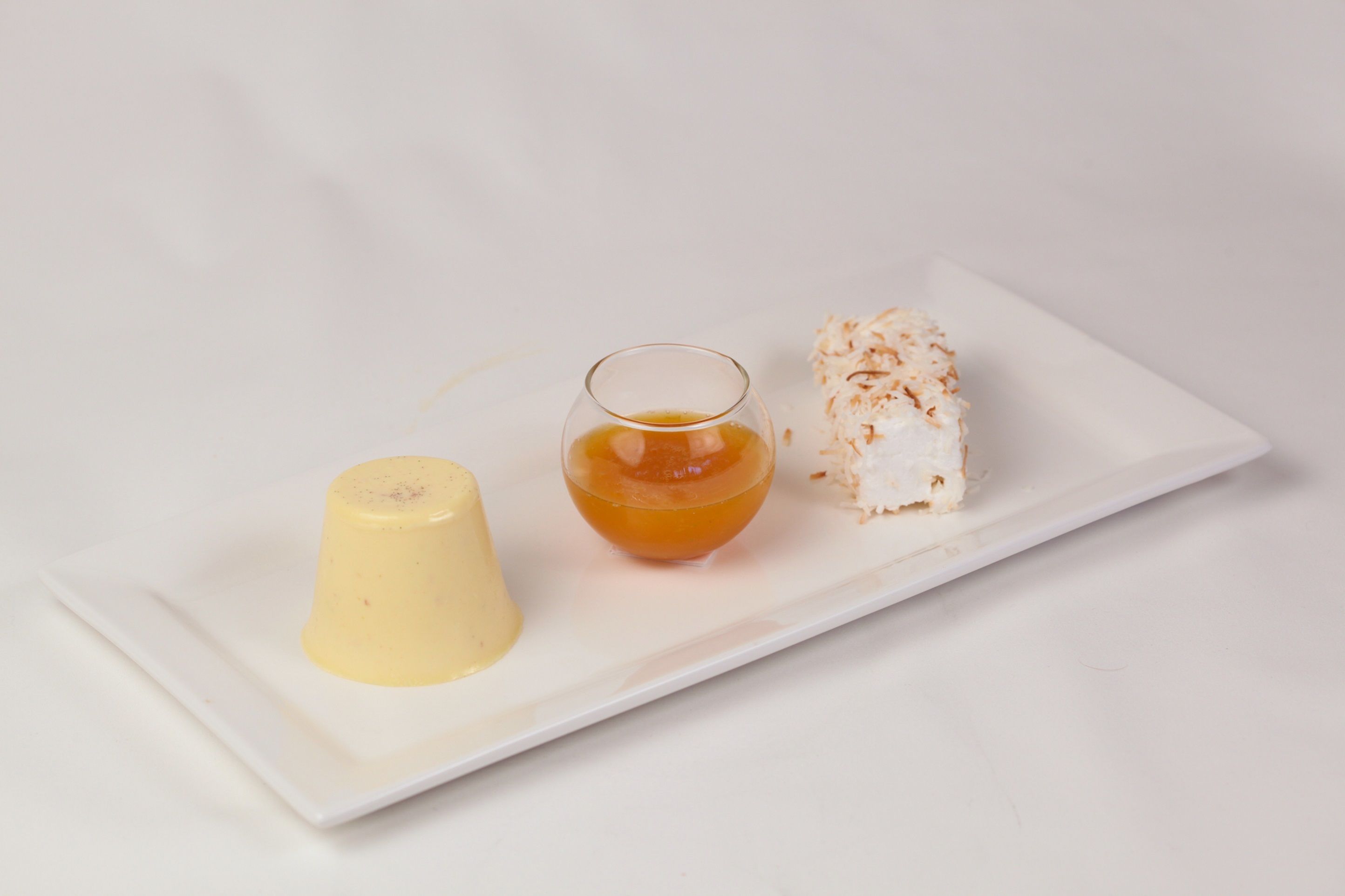 A dessert plate with a yellow mousse, a glass of orange liquid, and a white cake with coconut on a white plate.